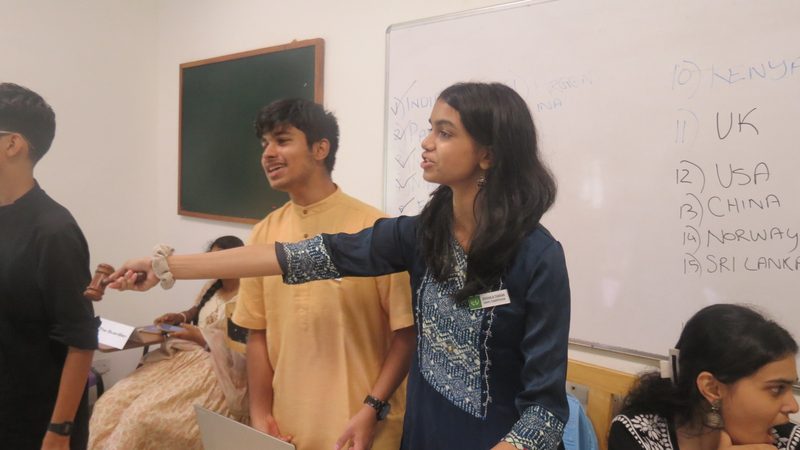 A delegate directs the floor, with the speakers list written on the board behind.